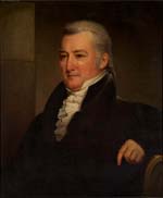 Seated, grey- haired white man, wearing a dark coat, ruffled white cravat, high collar, and with his elbow on the back of his chair. 